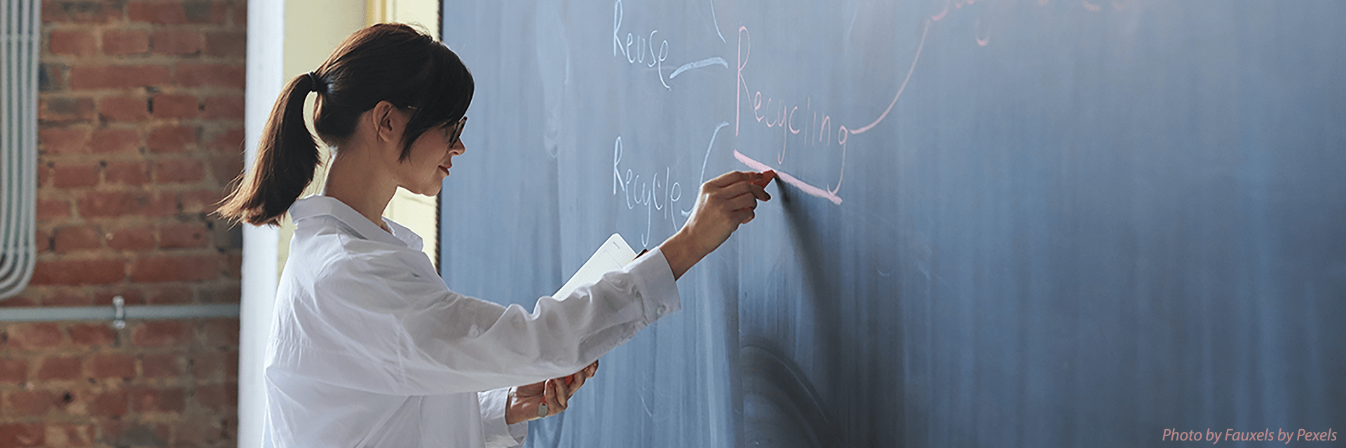 Femal instructor with her hair pulled back in a ponytail writes notes on a chalkboard as she teaches a class.